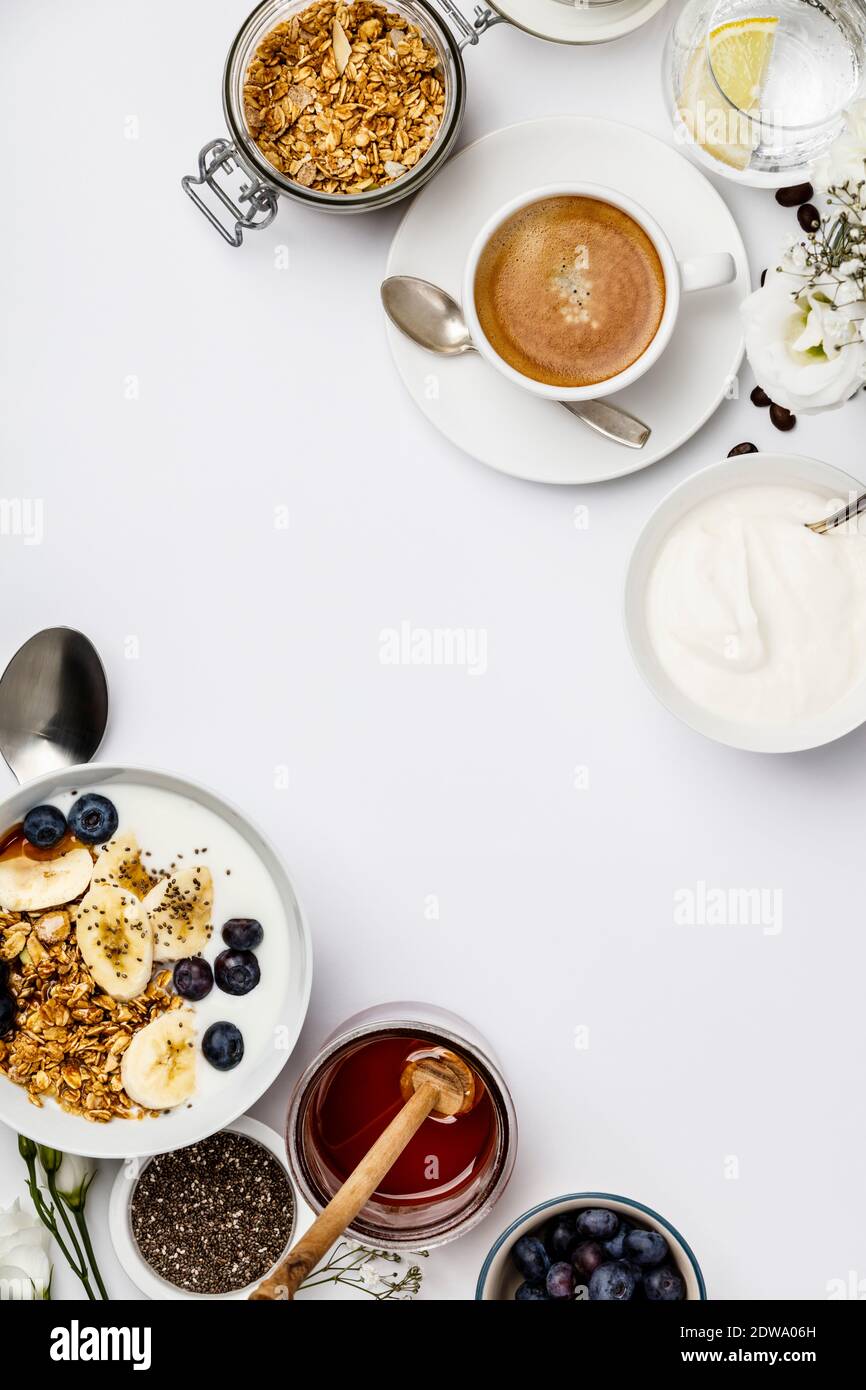 Gesundes Frühstücksset. Hafergranola mit Joghurt, Honig, frischen Bananen, Heidelbeeren, Chiasamen in Schüssel und Tasse Kaffee auf weißem Hintergrund, Draufsicht, Stockfoto