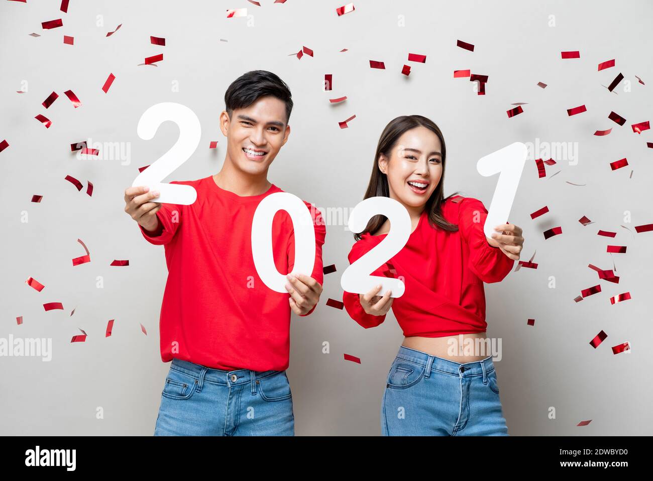 Lächelndes glückliches asiatisches Paar in roter legerer Kleidung mit 2021 Zahlen für das Jahreskonzept auf hellgrauem Hintergrund mit Konfetti Stockfoto