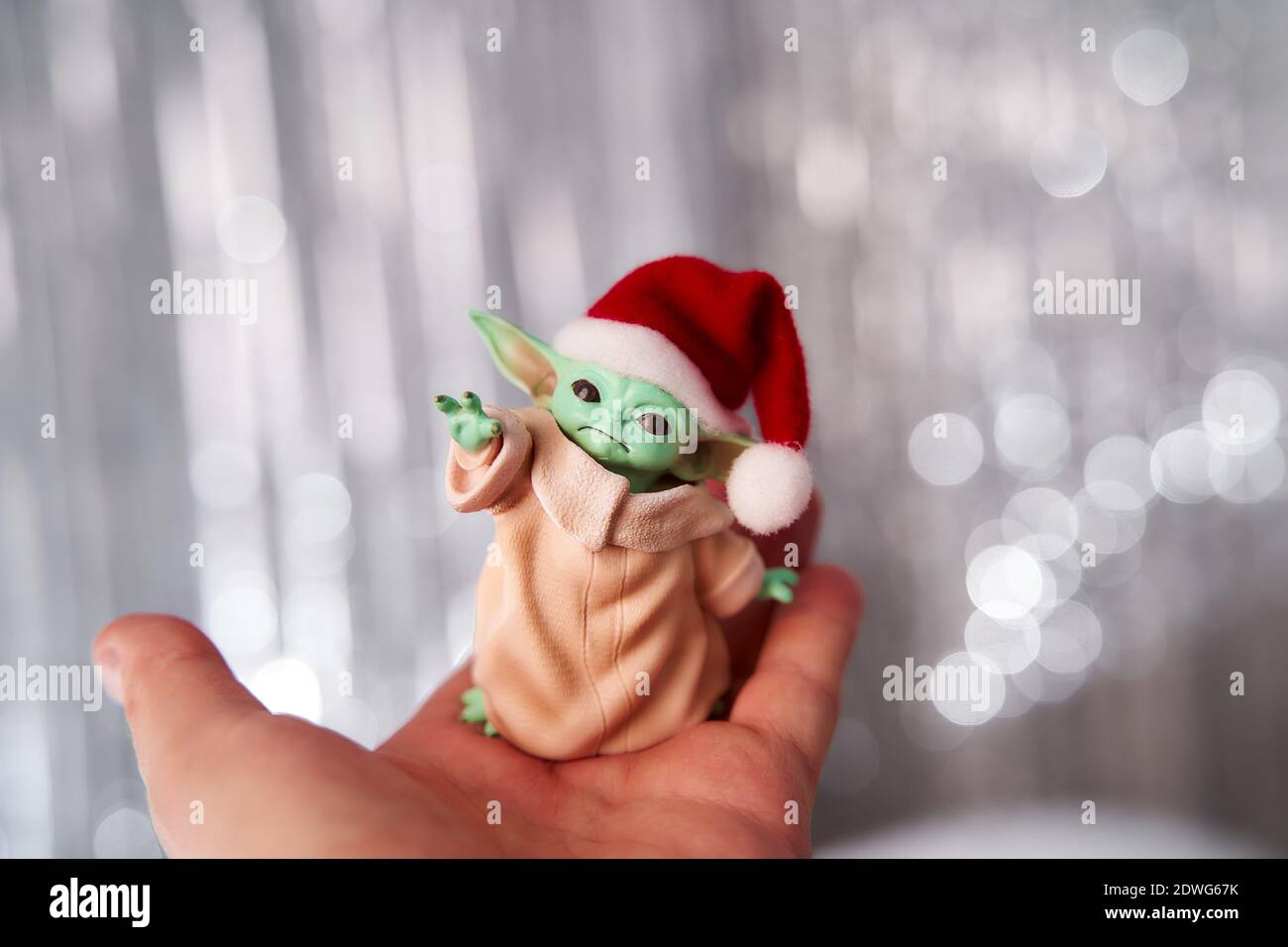 Dez, 2020: Darstellung von Baby Yoda, einer Action-Figur, die in einer roten Mütze auf weihnachtlichem Hintergrund steht. Bokeh-Effekt. Stockfoto