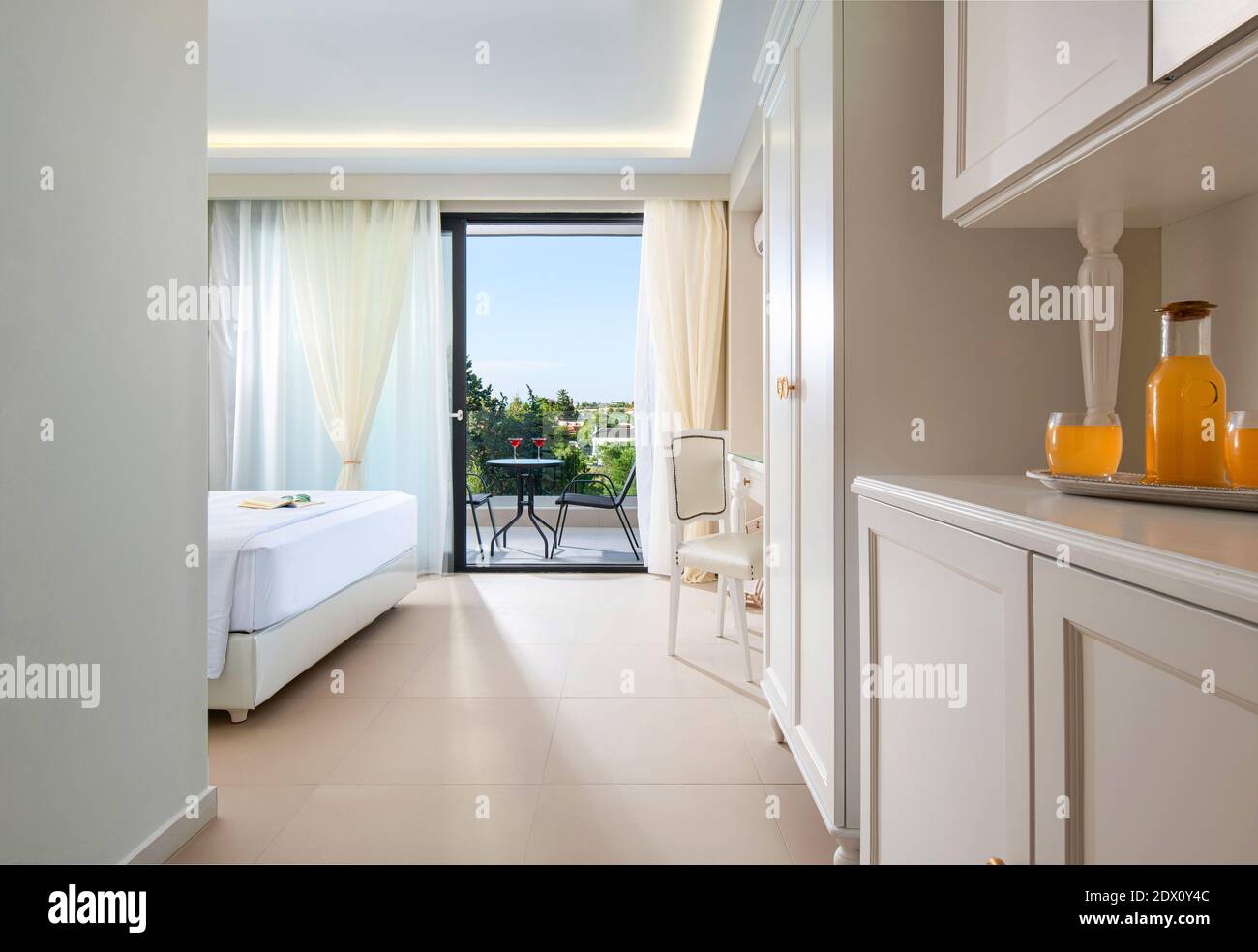 Entspannungskonzept des klassischen Innenresort-Hotelapartments mit einem Begrüßungsgetränk mit Orangensaft. Weißes Schlafzimmer, großes, offenes Fenster - Glastür, um es zusammenzufassen Stockfoto