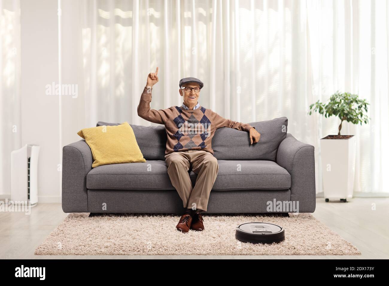 Lächelnder älterer Mann, der zu Hause auf einem Sofa sitzt, nach oben zeigt und ein Roboter-Staubsauger den Teppich staubt Stockfoto