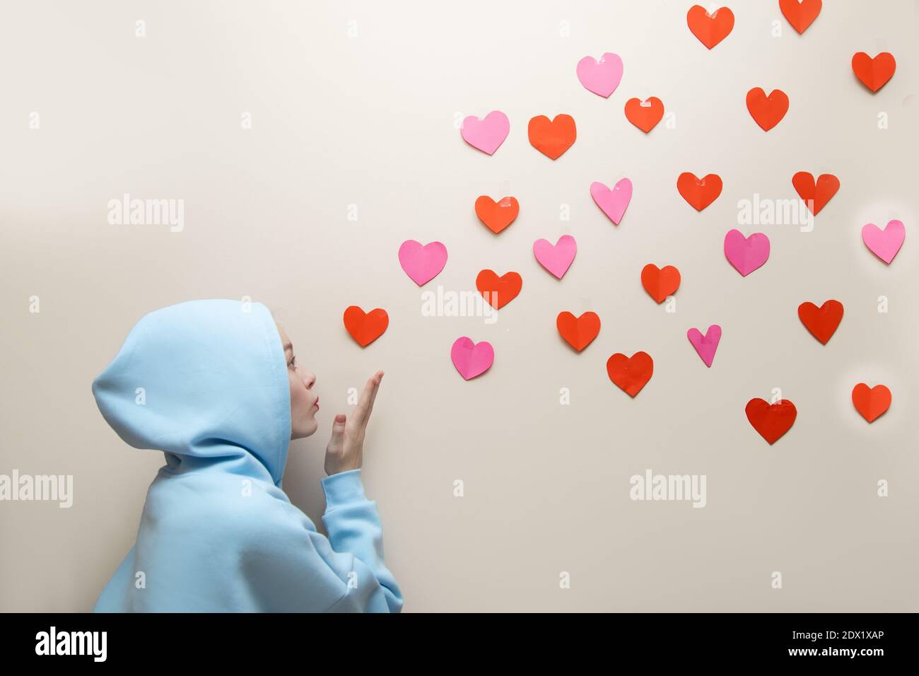 Young Girl im blauen Hoodie mit Kapuze bläst Herzen von ihrer Handfläche und sie fliegen entlang der Wand, kopieren Raum. Valentinstag, Suche Liebe Konzept, geben g Stockfoto