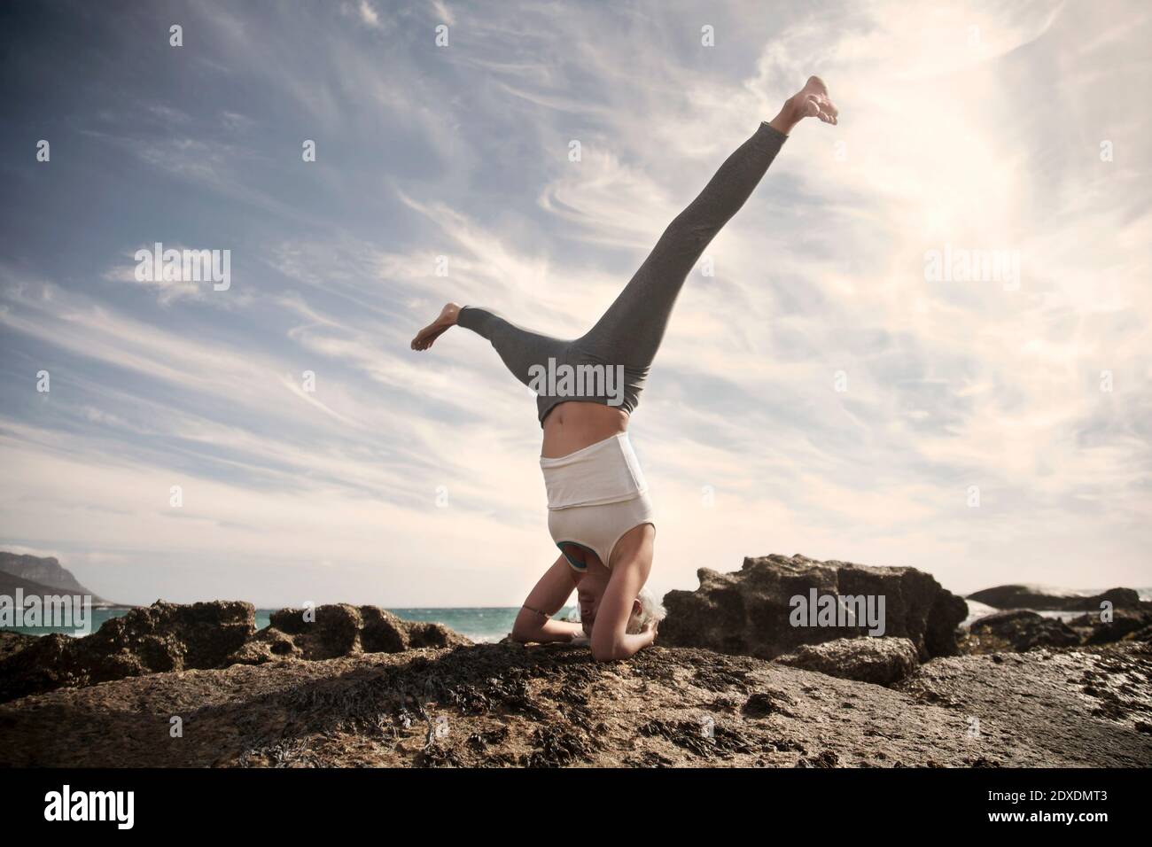 Sorglose Frau, die Sirsasana mit gespreizten Beinen auf Felsformation praktiziert Am Strand gegen Himmel Stockfoto
