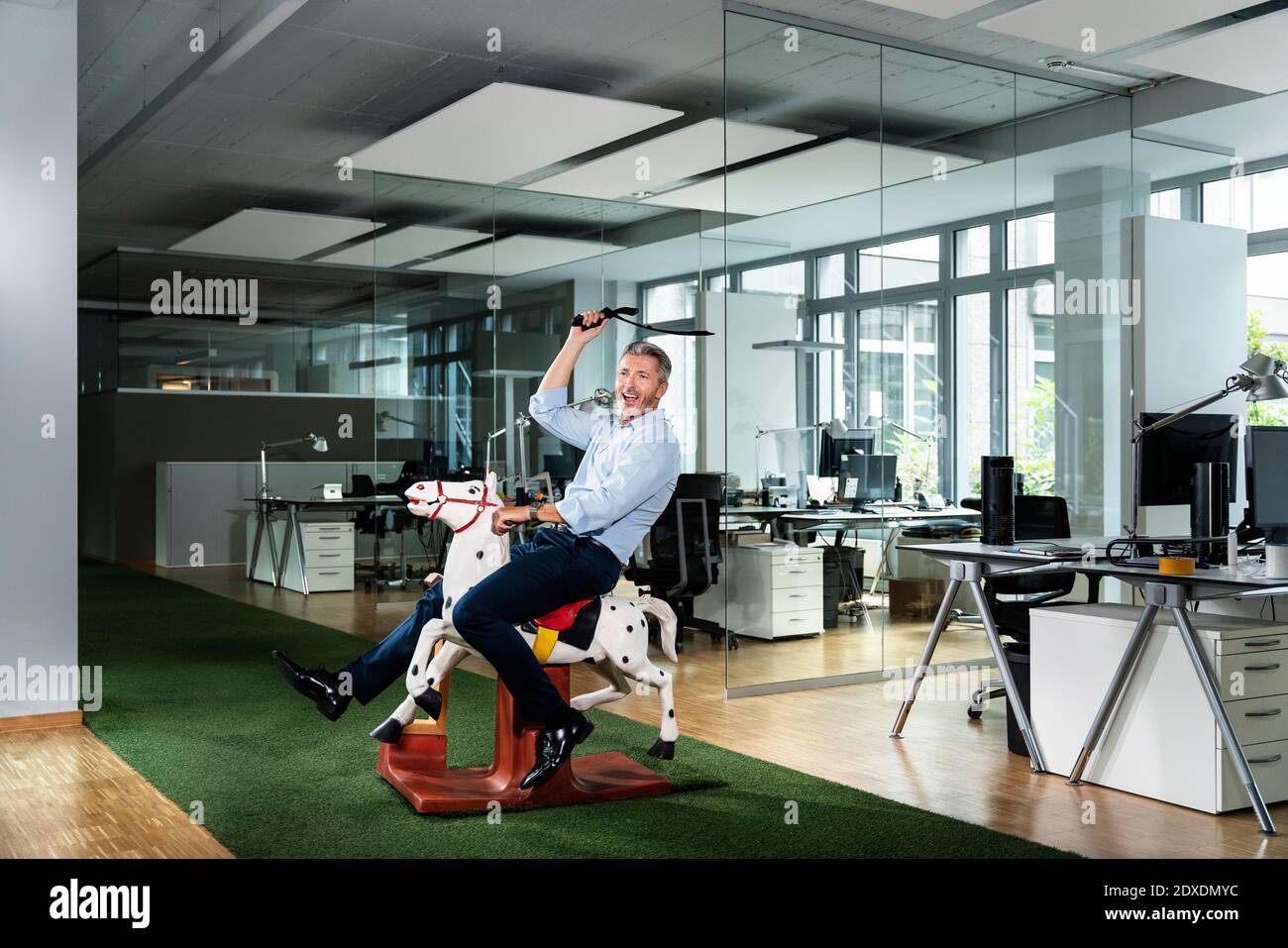 Fröhlicher Geschäftsmann schaut weg, während er auf Pferd Spielzeug sitzt Büro Stockfoto