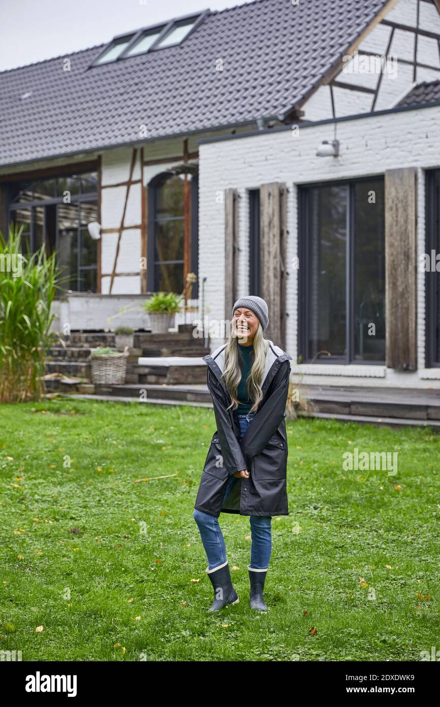 Lachende reife Frau im Regenmantel, die auf Gras gegen Haus steht Stockfoto