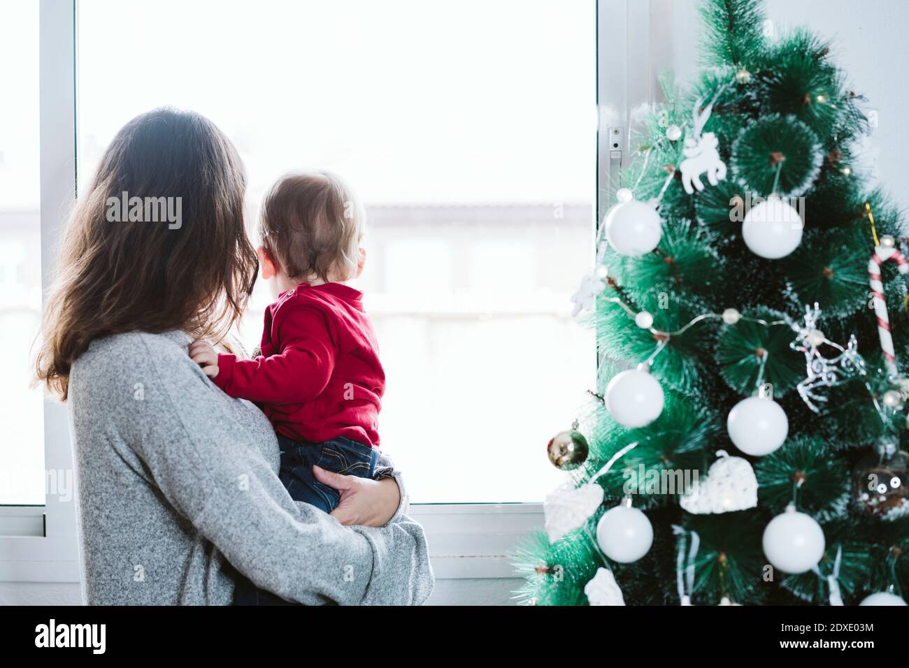 Mutter mit Baby Junge, der durch das Fenster schaut, während er ansteht Zuhause während Weihnachten Stockfoto