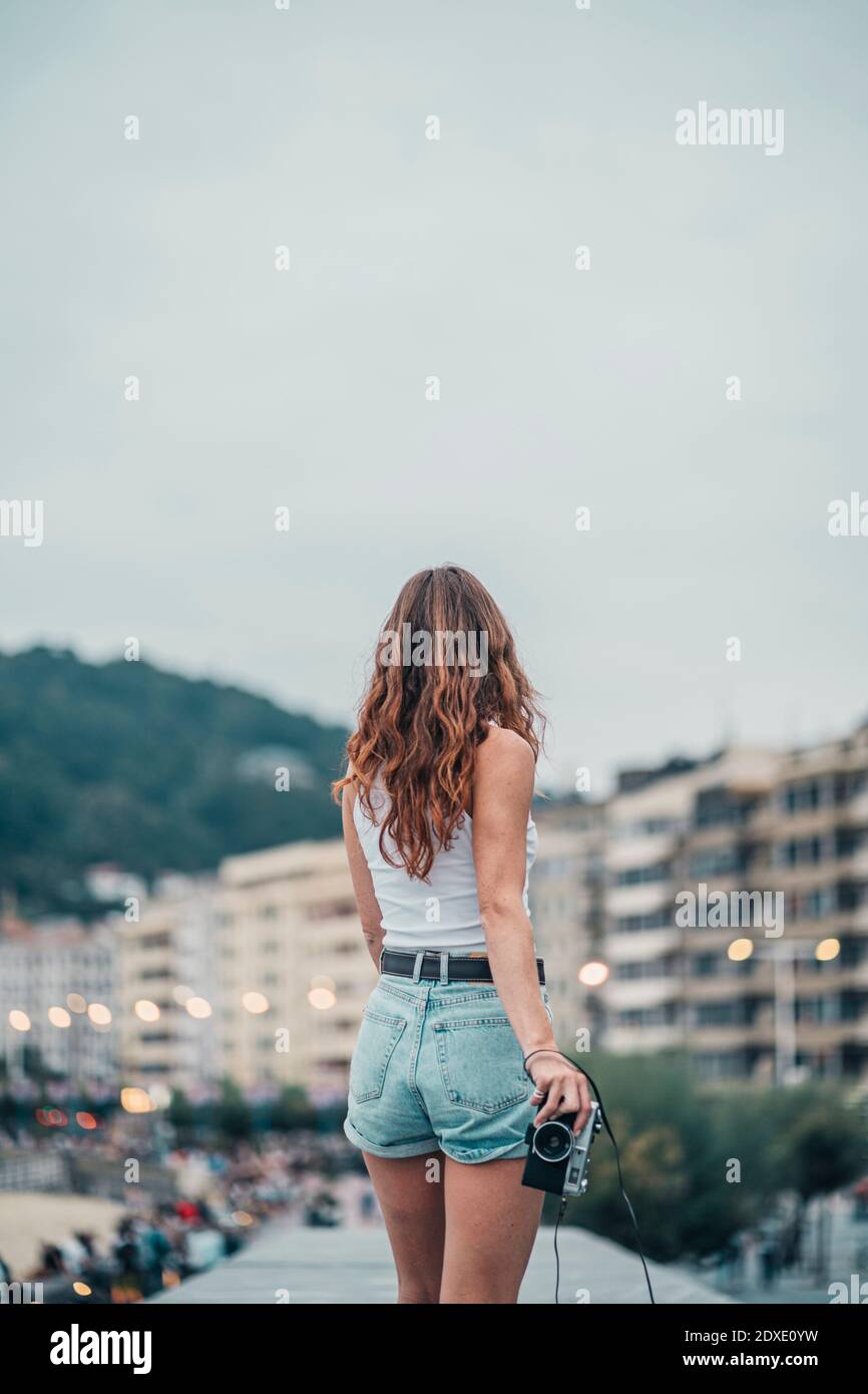 Junge Frau mit Kamera, die im Stehen auf die Stadt blickt Gegen den Himmel Stockfoto