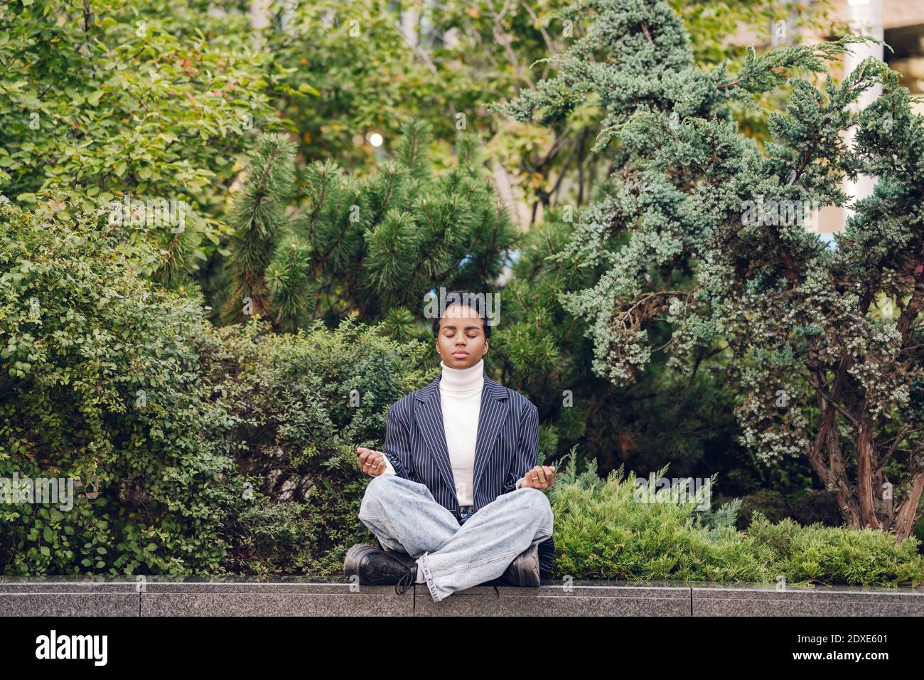 Junge Geschäftsfrau, die Yoga macht, während sie auf einer Stützmauer sitzt Anlagen Stockfoto