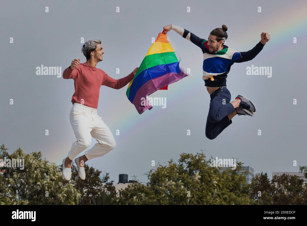 Lächelnde Männer mit Schal springen gegen Regenbogen in klarem Himmel Stockfoto