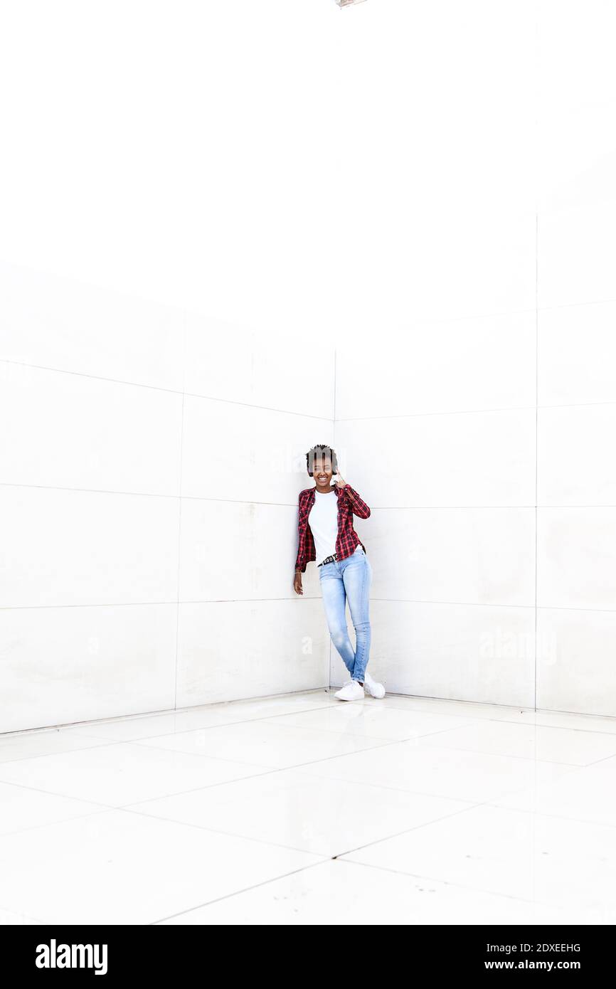 Lächelnde junge Frau, die sich an der weißen Wand mit gekreuzten Beinen lehnt Stockfoto