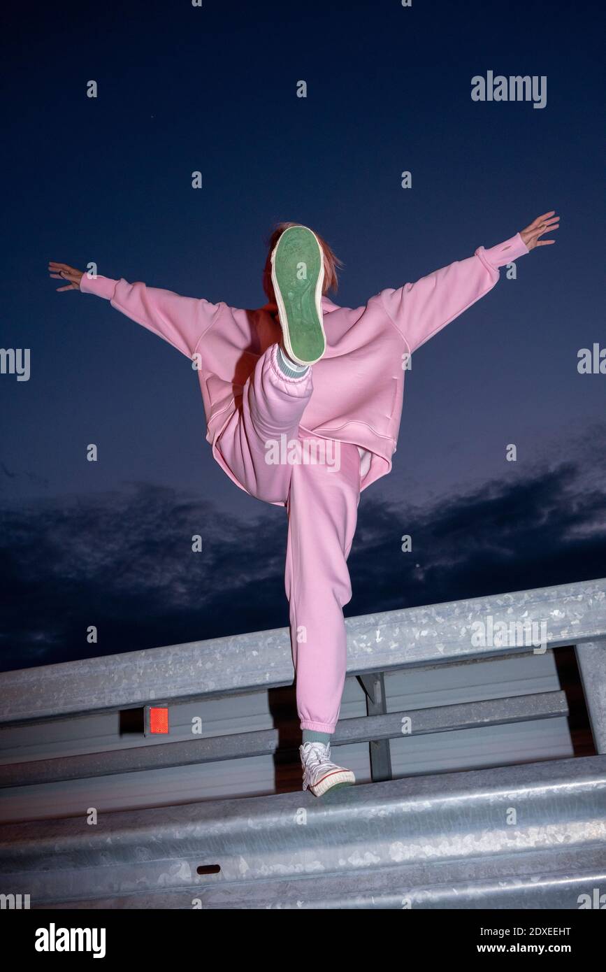 Junge Frau in pinkem Anzug, der auf einem Bein steht Nachts an der Straßenbarriere Stockfoto