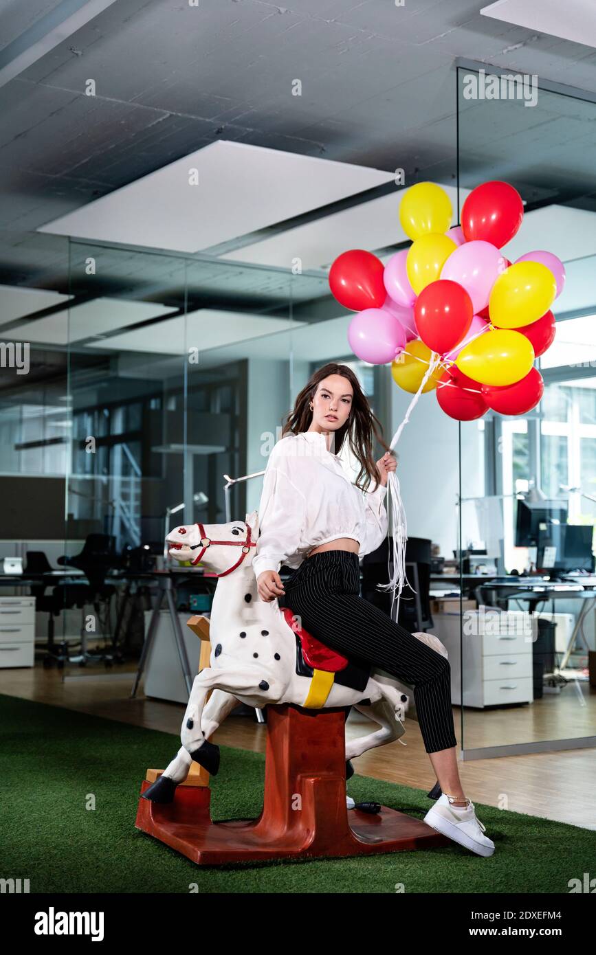 Junge Geschäftsfrau mit mehrfarbigem Ballon sitzt auf Schaukelpferd Im Büro Stockfoto