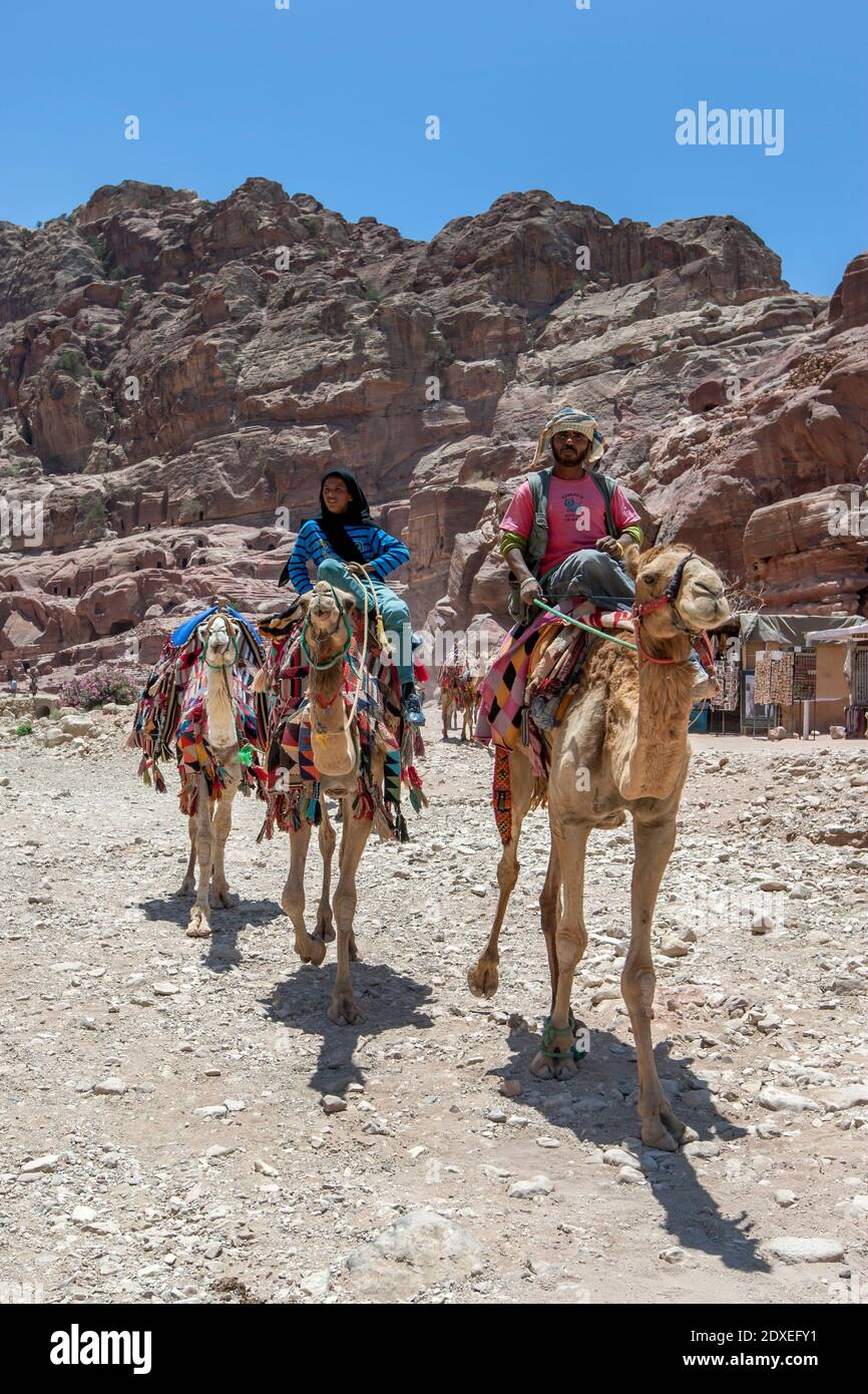 Beduinenmänner reiten auf Kamelen mit bunten Teppichen durch die antike Stätte Petra in Jordanien. Sie nehmen Touristen für Fahrten durch die Ruinen von Petra. Stockfoto