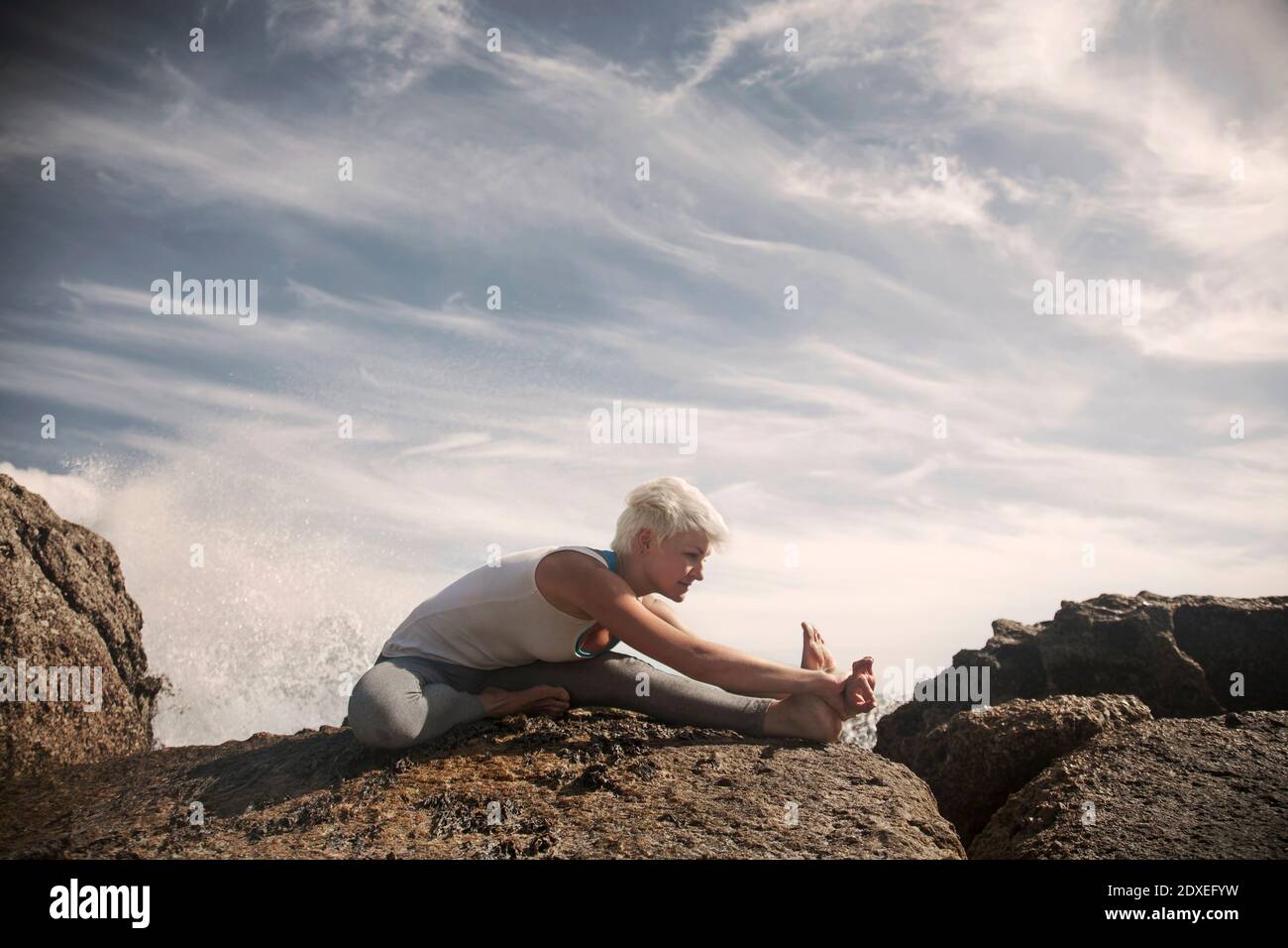 Blonde flexible Frau praktiziert Yoga auf Felsformation am Strand Gegen den Himmel Stockfoto