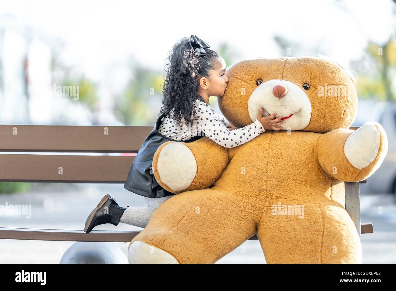 Mädchen küssen Teddybär beim Knien auf Bank Stockfoto