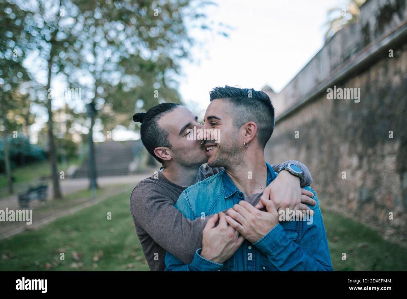Glücklicher Mann küsst Freund auf Wange, während umarmt von hinten Im öffentlichen Park Stockfoto