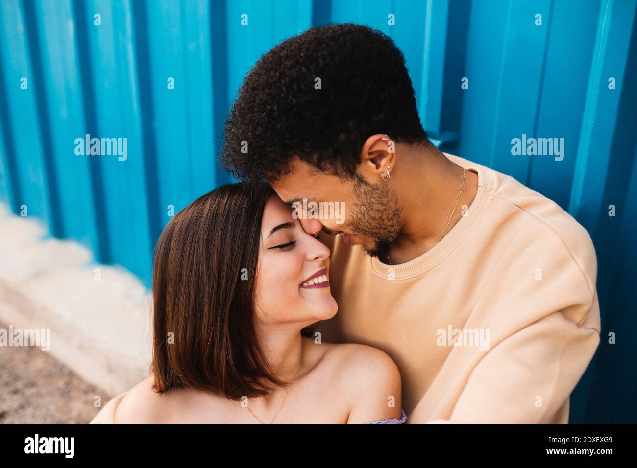 Lächelndes junges Paar, das von Angesicht zu Angesicht gegen die blaue Wand sitzt Stockfoto