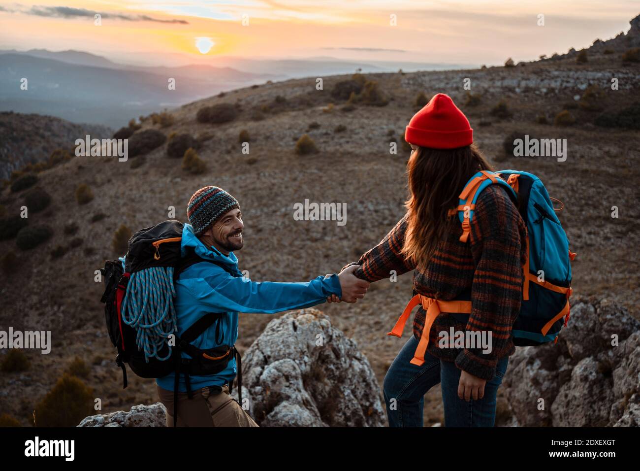 Freundin hilft Freund zu klettern felsigen Berg während Sonnenuntergang Stockfoto