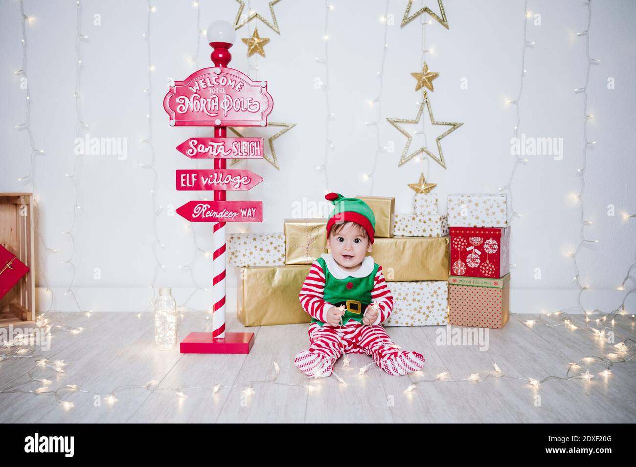 Kleiner Junge im Elf Kostüm, der mit Lichtern spielt, während er sitzt Mit Willkommensschild zu Hause während Weihnachten Stockfoto