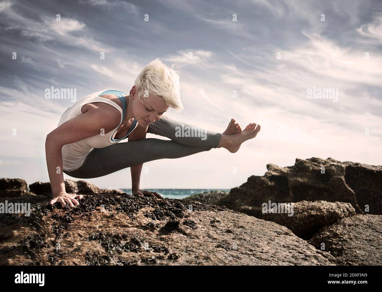 Blonde Frau üben Astavakrasana auf Felsformation am Strand gegen Himmel Stockfoto
