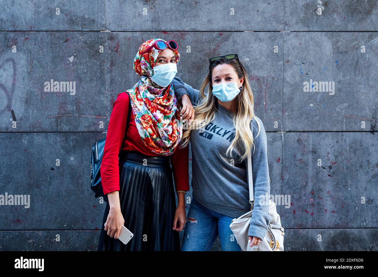 Frau trägt Gesichtsmaske, während sie mit der Hand auf Muslim steht Die Schulter eines Freundes gegen die graue Wand während einer Pandemie Stockfoto