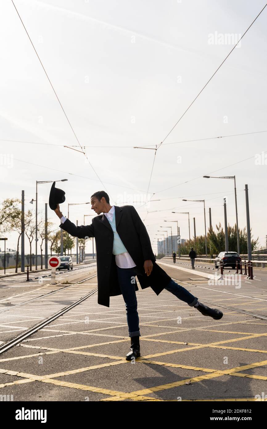 Junger Mann, der auf einem Bein steht und dabei den Hut zwischen sich hält Eisenbahnstrecke auf der Straße Stockfoto
