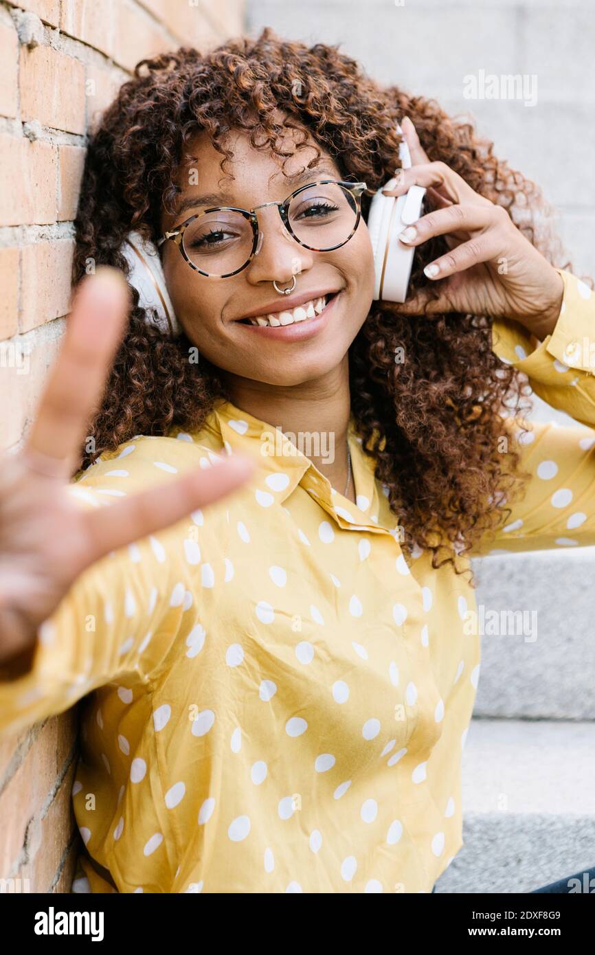 Glückliche Frau mit Kopfhörern, die während des Sitzens eine Friedensgeste zeigt Schritte Stockfoto