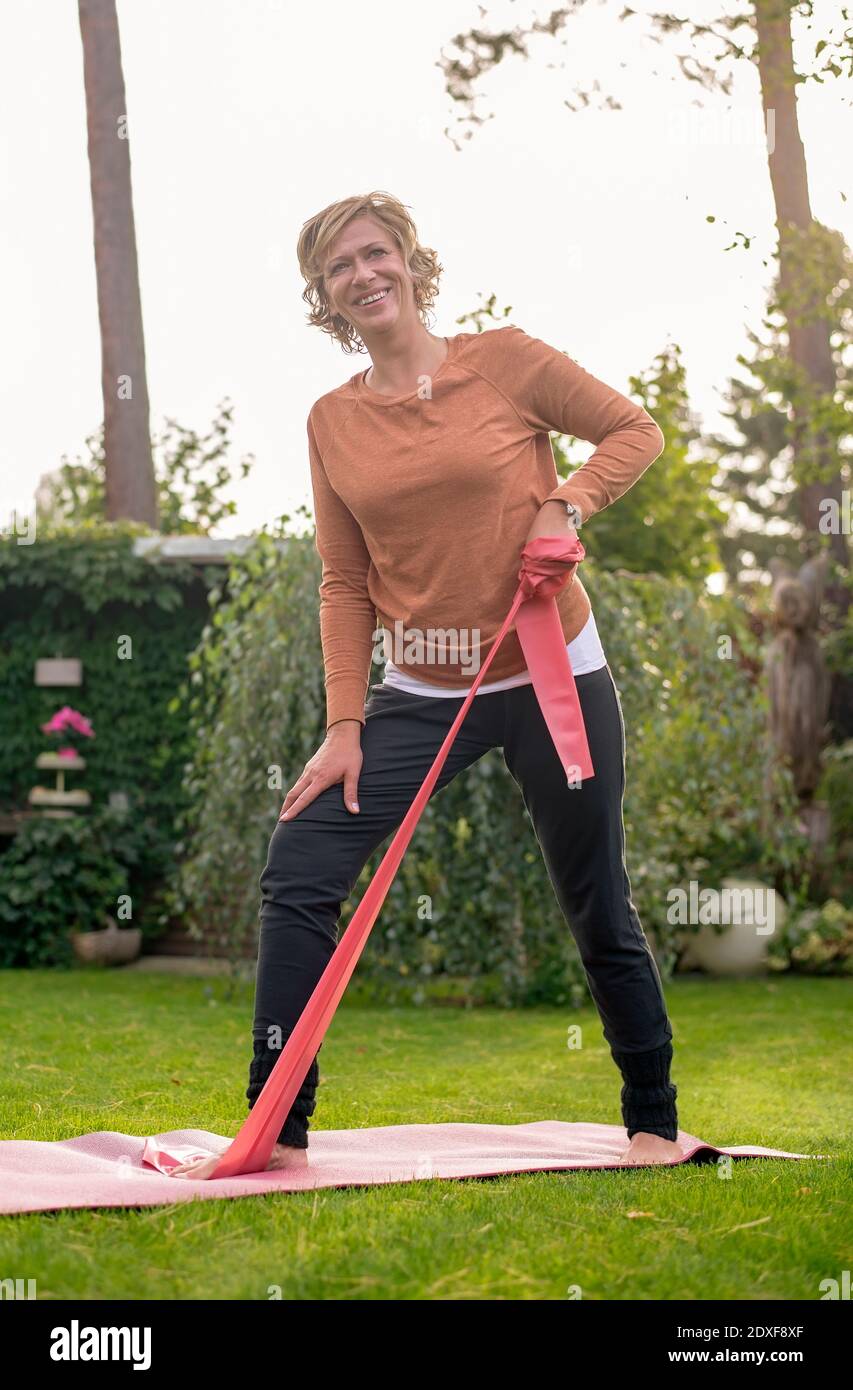Lächelnde Frau, die mit Widerstandsband trainiert, während sie auf dem Rücken steht Hof Stockfoto