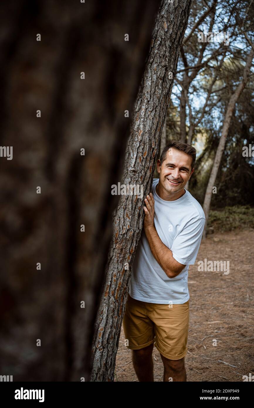Lächelnder Mann, der während des Urlaubs am Baumstamm im Wald steht Stockfoto