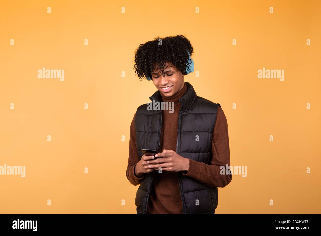 Foto von jungen Mann in dunkler Weste Telefon halten, Freunde chatten, Musik moderne Technologie mit blauen Kopfhörern. Isoliertes Bild auf gelbem Hintergrund. Stockfoto