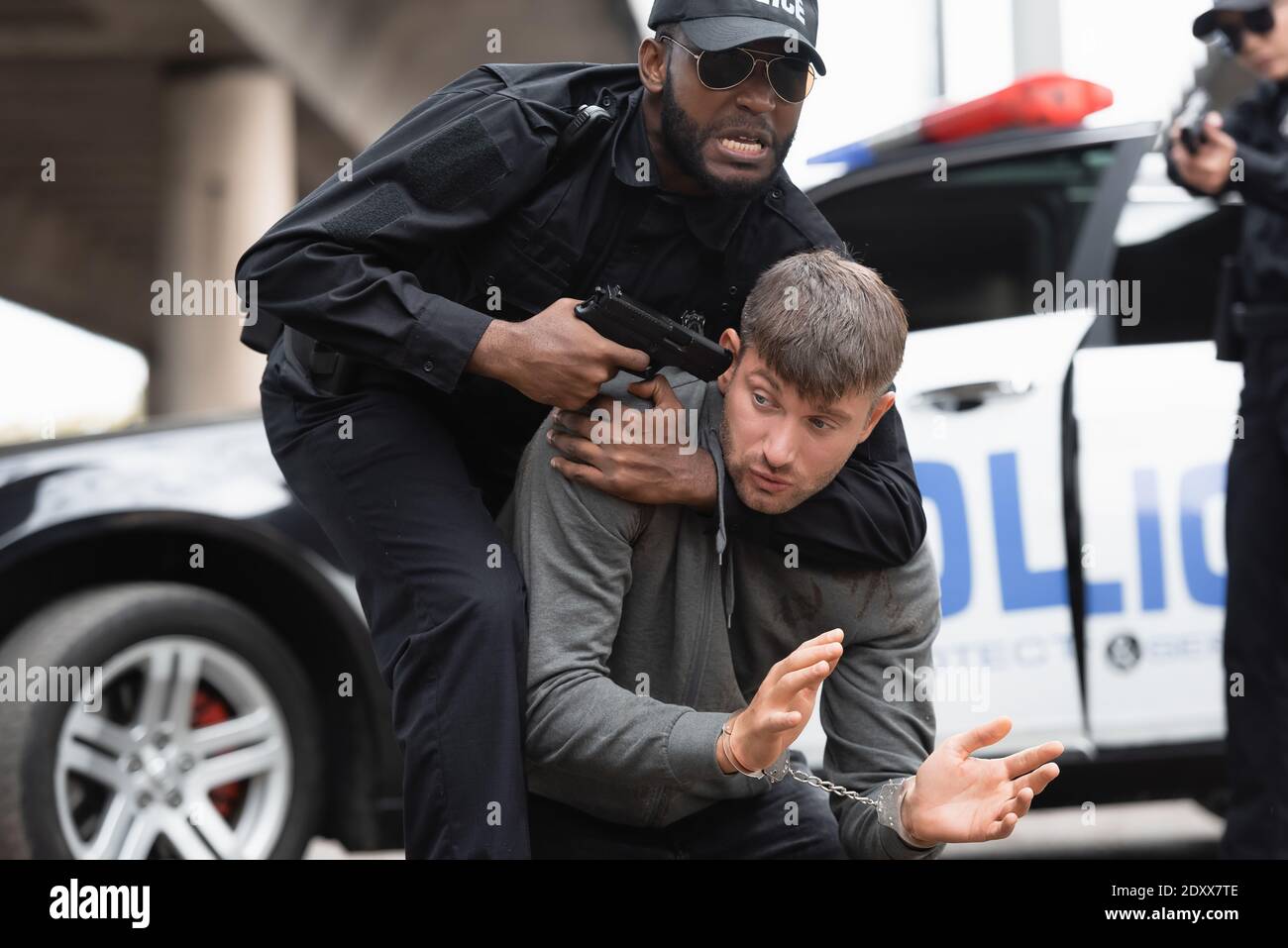 Wütender afroamerikanischer Polizist, der mit Pistole auf einen mit Handschellen gefesselten Täter zielt Bei unscharfem Hintergrund im Freien Stockfoto