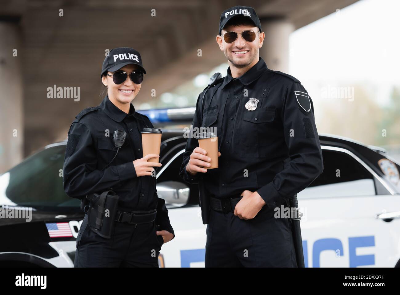 Lächelnde Polizisten in Sonnenbrillen halten Kaffee zum Mitnehmen in der Nähe des Autos Bei unscharfem Hintergrund im Freien Stockfoto