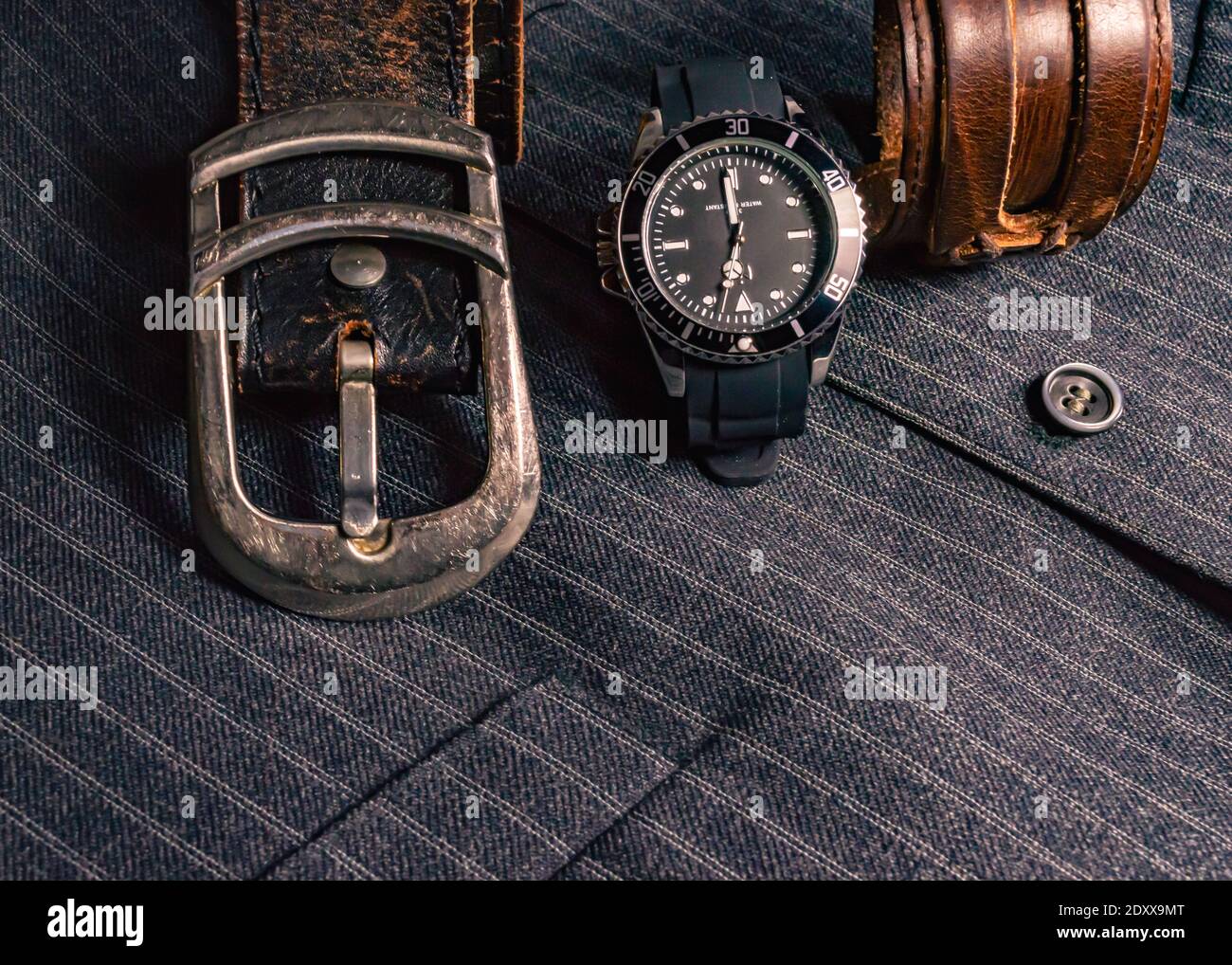 Abbildung eines Ledergürtels, einer Uhr in Schwarz und Chrom und einem Lederarmband auf einer grau gestreiften Weste Stockfoto