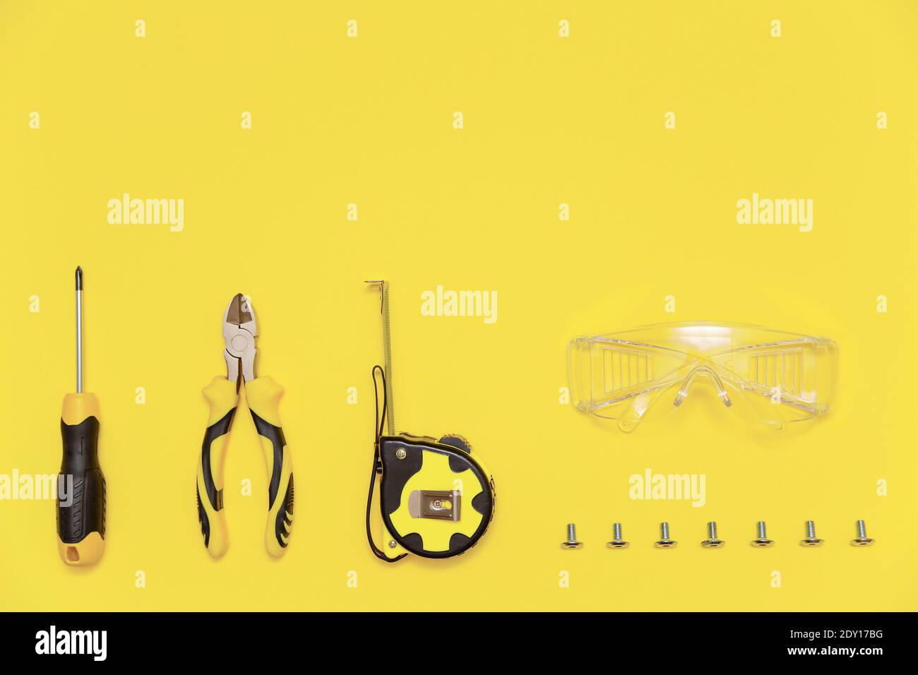 Eine Flachlage mit einem Satz von Tischwerkzeugen, Schraubendreher, Schneidzange, Maßband, Schutzbrille und Schrauben auf gelbem beleuchtenden Hintergrund Stockfoto