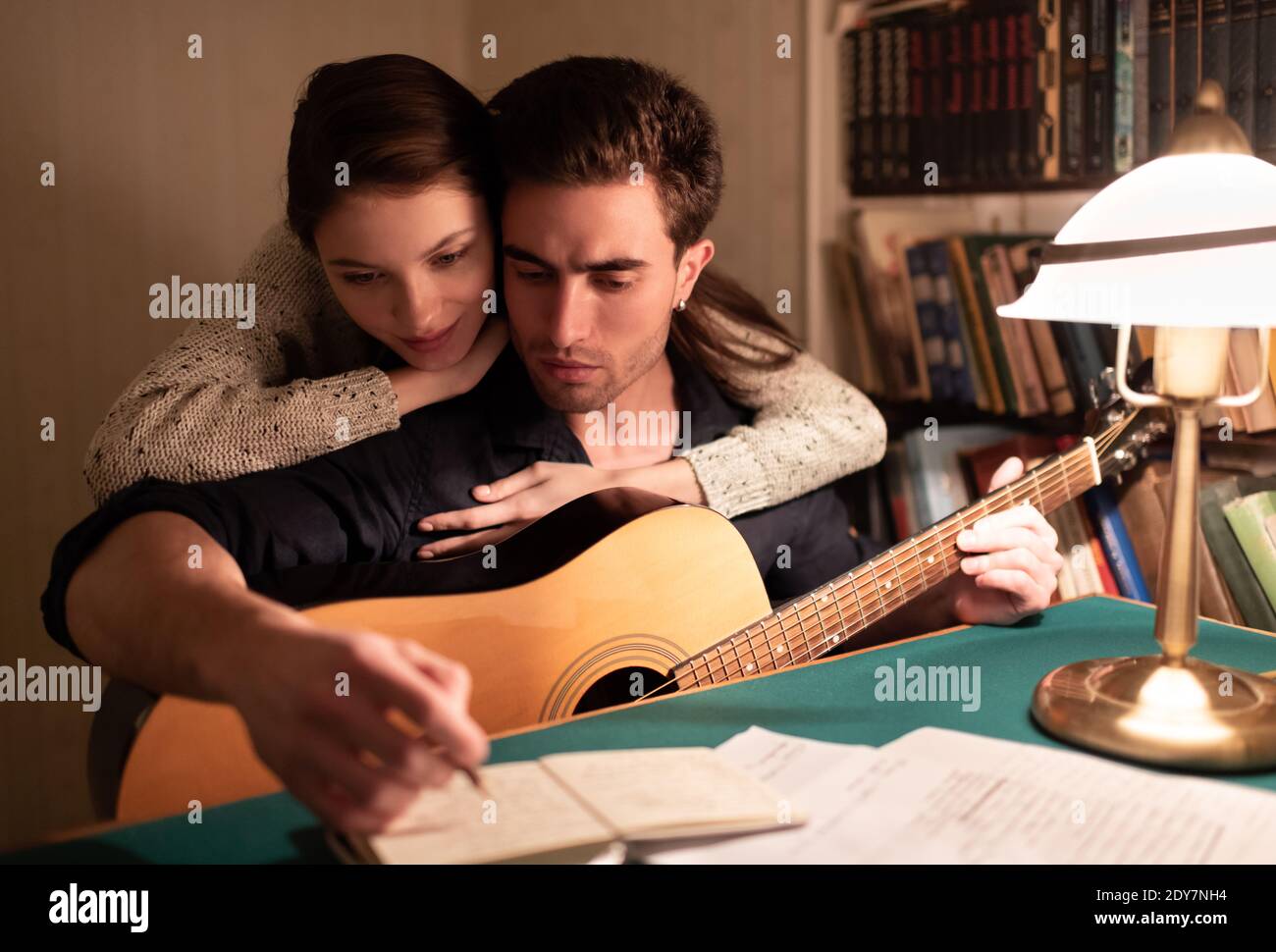 Junge Frau umarmt Freund beim Lesen von Notizen in Notizblock während Prozess der Songerstellung in der alten Wohnung am Abend Stockfoto