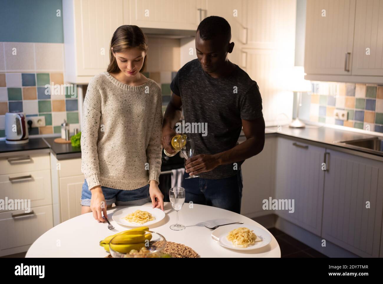 Afroamerikanischer Mann filing Gläser mit Wein, während junge Frau Putting Teller mit Spaghetti auf dem Tisch während romantische Datum an Zu Hause Stockfoto