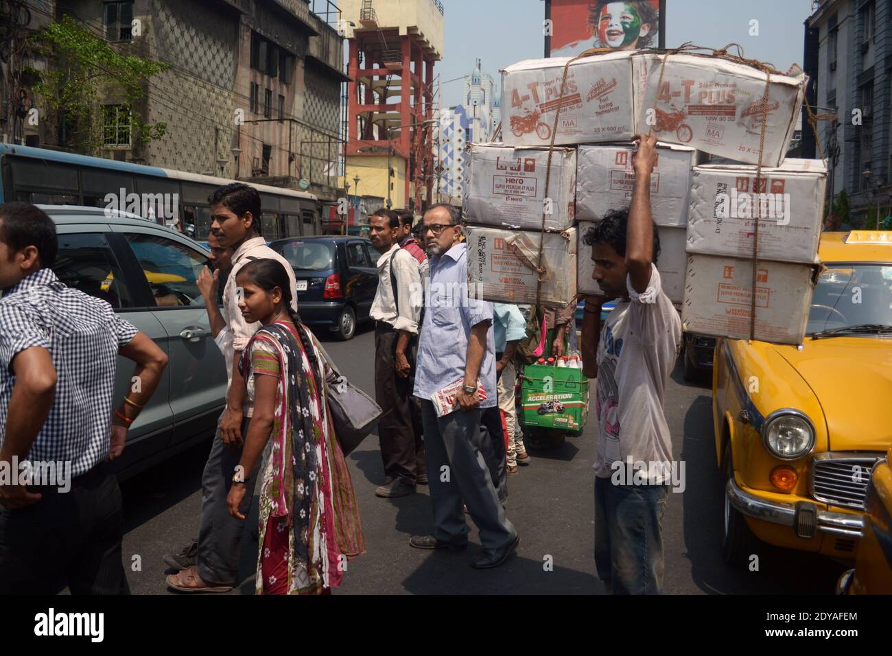 Kolkata, Indien - März, 2014: Mann, der schwere Kisten trägt und Straße mit anderen Leuten auf Fußgänger kreuzt. Indischer Stadtverkehr in Kalkutta Stockfoto