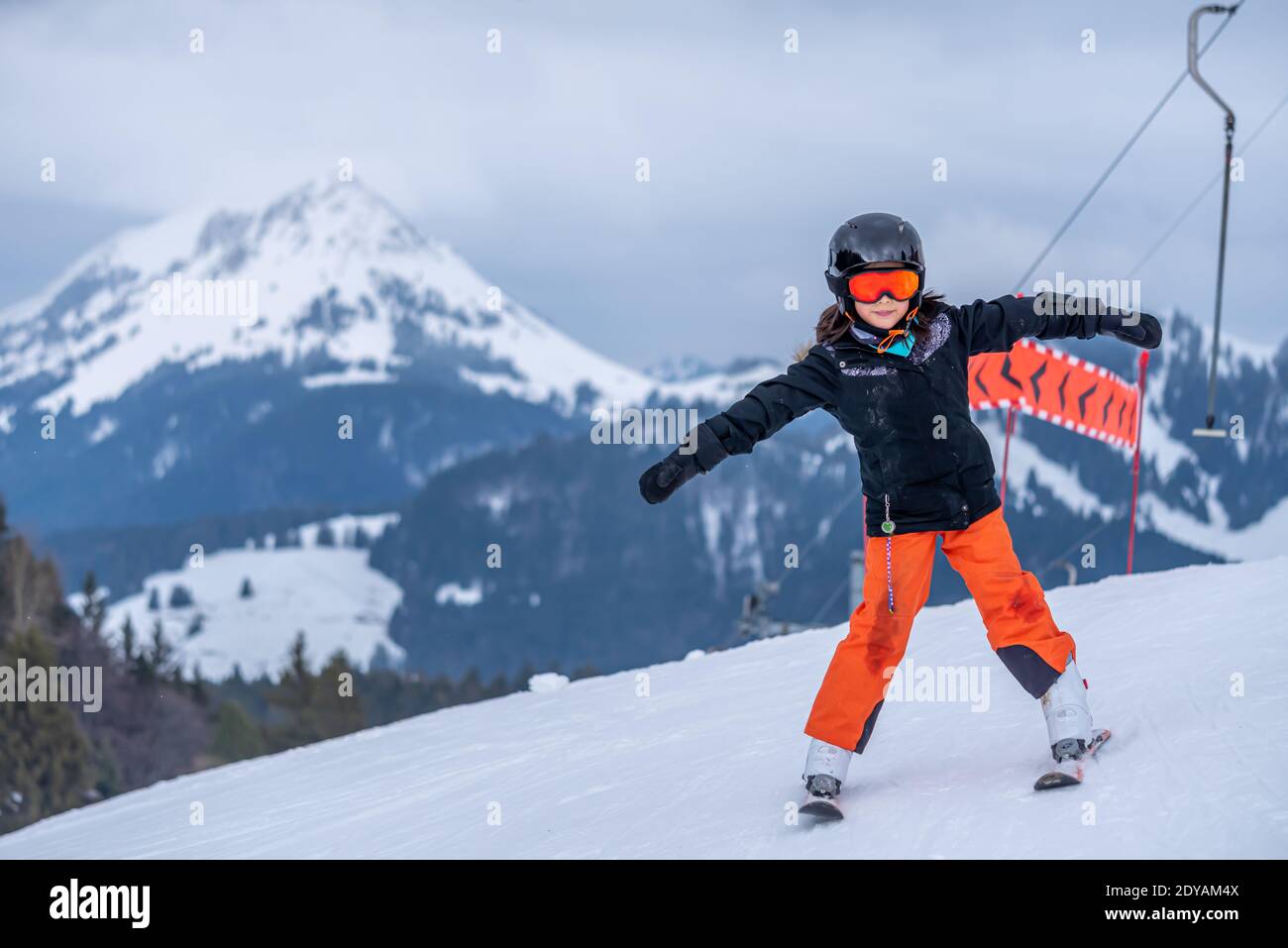 Skifahren im Schnee. Ein asiatisches Mädchen, das auf dem Skilift sitzt, indem es das Seil ergreifet und Skistöcke hält. Urlaub in der Schweiz. Frohe Kindheit. Stockfoto