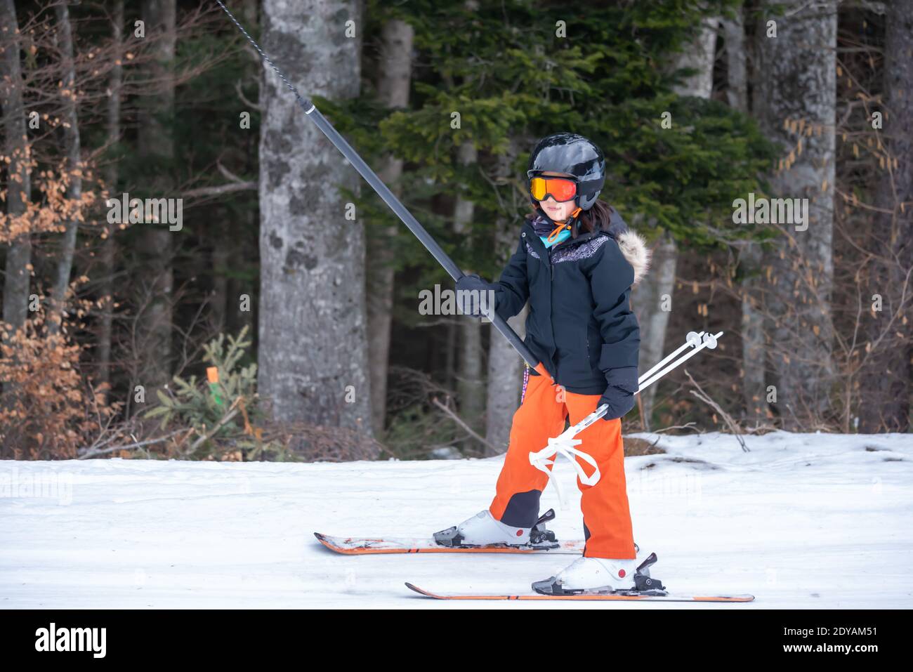 Skifahren im Schnee. Ein asiatisches Mädchen, das auf dem Skilift sitzt, indem es das Seil ergreifet und Skistöcke hält. Urlaub in der Schweiz. Glückliche Kindheit, Stockfoto