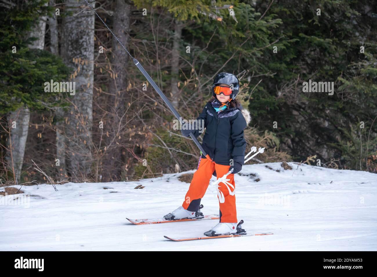 Skifahren im Schnee. Ein asiatisches Mädchen, das auf dem Skilift sitzt, indem es das Seil ergreifet und Skistöcke hält. Urlaub in der Schweiz. Glückliche Kindheit, Stockfoto