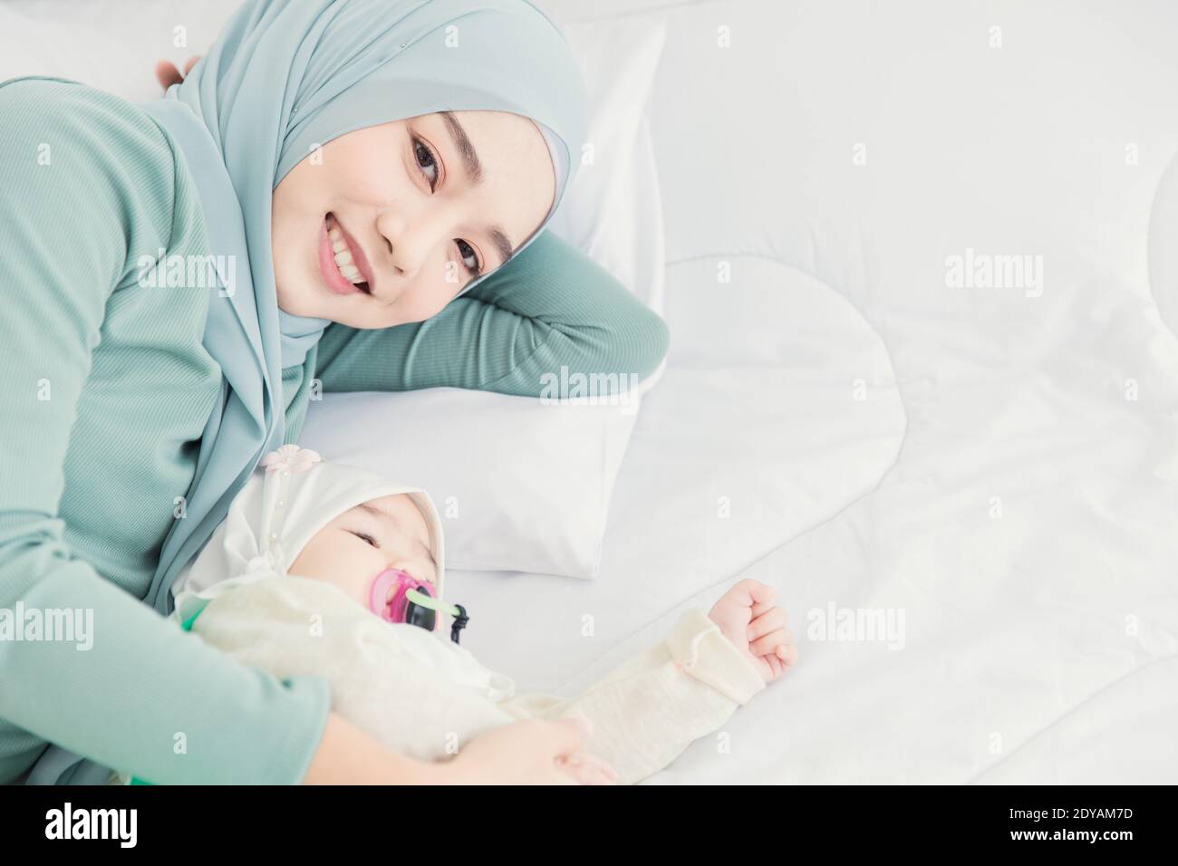 Muslim Hijab Aufenthalt mit ihrem Baby auf weißem Bett lächelnd zu Hause, Mutter Pflege Kleinkind. Stockfoto