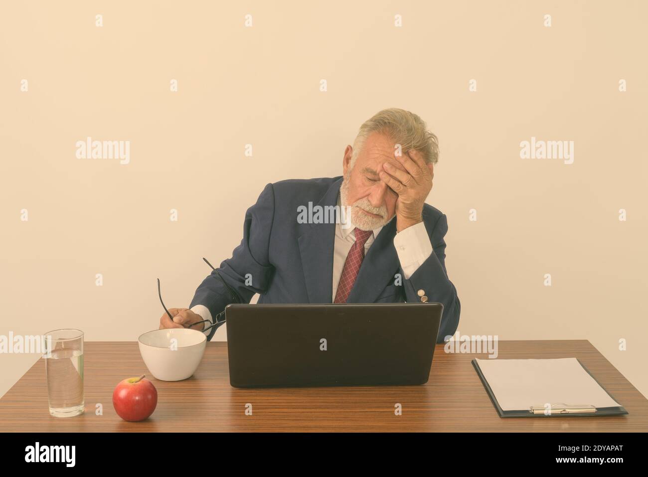 Studioaufnahme eines gutaussehenden älteren bärtigen Geschäftsmannes, der während der Aufnahme eine Brille hält Kopfschmerzen mit Laptop und grundlegende Dinge für die Arbeit an Holztisch Stockfoto