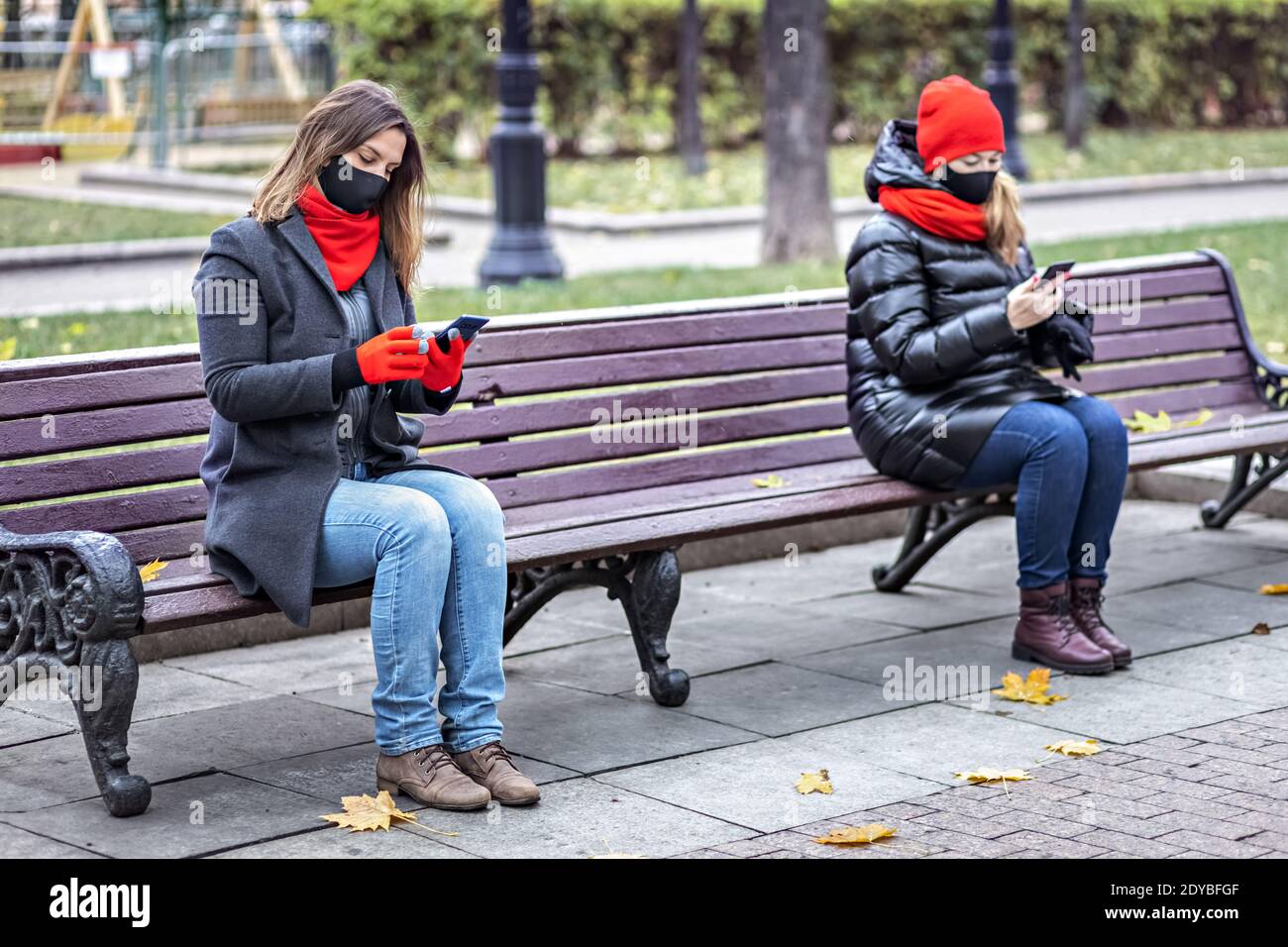 Zwei junge Frauen mit medizinischen Masken sprechen auf einem Handy in sicherer sozialer Distanz, während sie auf einer Bank in einem Herbstpark sitzen. Meeting und chatti Stockfoto