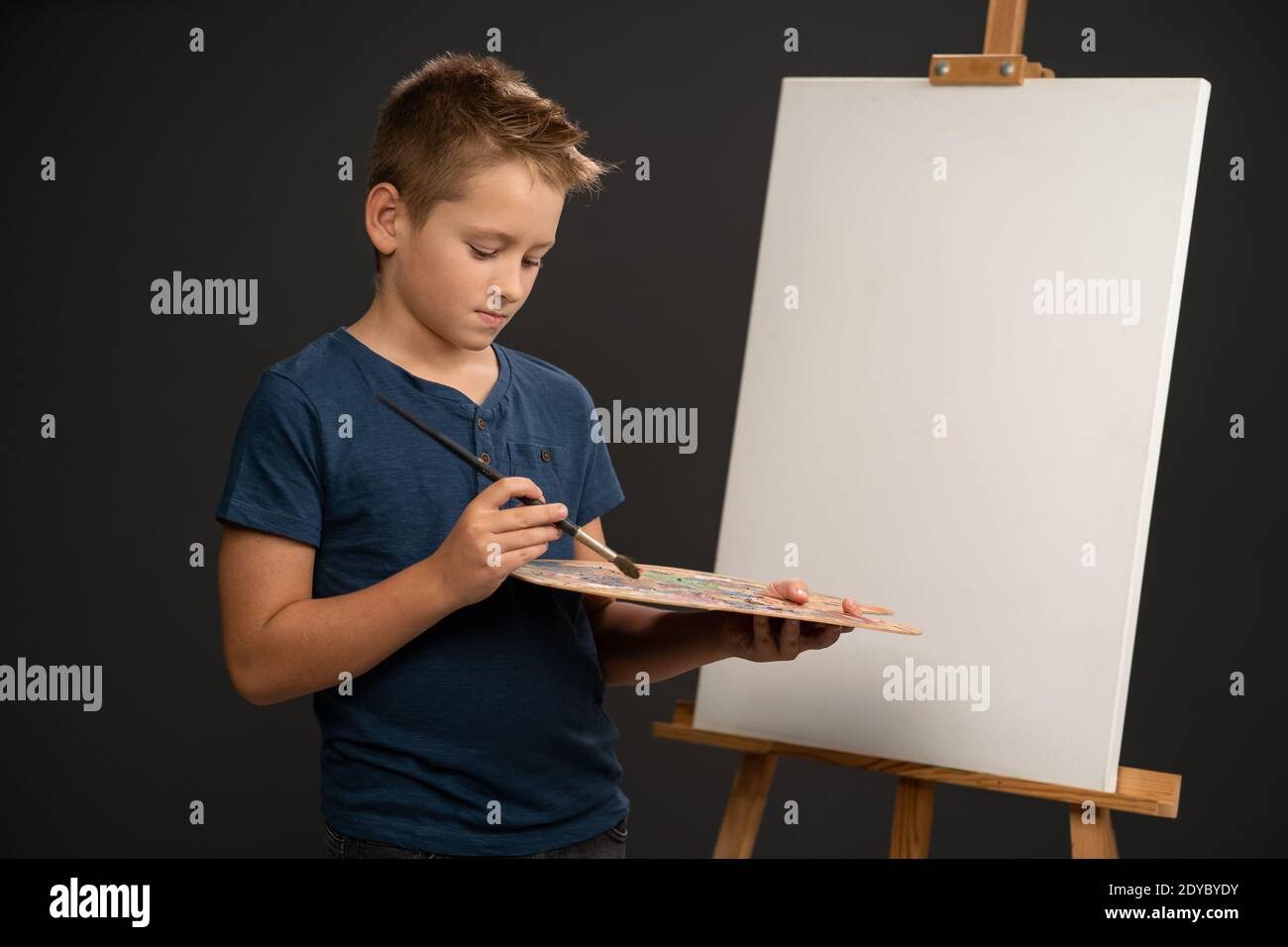 Mischen Farben bekommen die gewünschte Farbe halten eine Palette mit Malt 10 Jahre alten Jungen in blau T-Shirt sieht an Kamera auf dem Hintergrund einer Staffelei Stockfoto