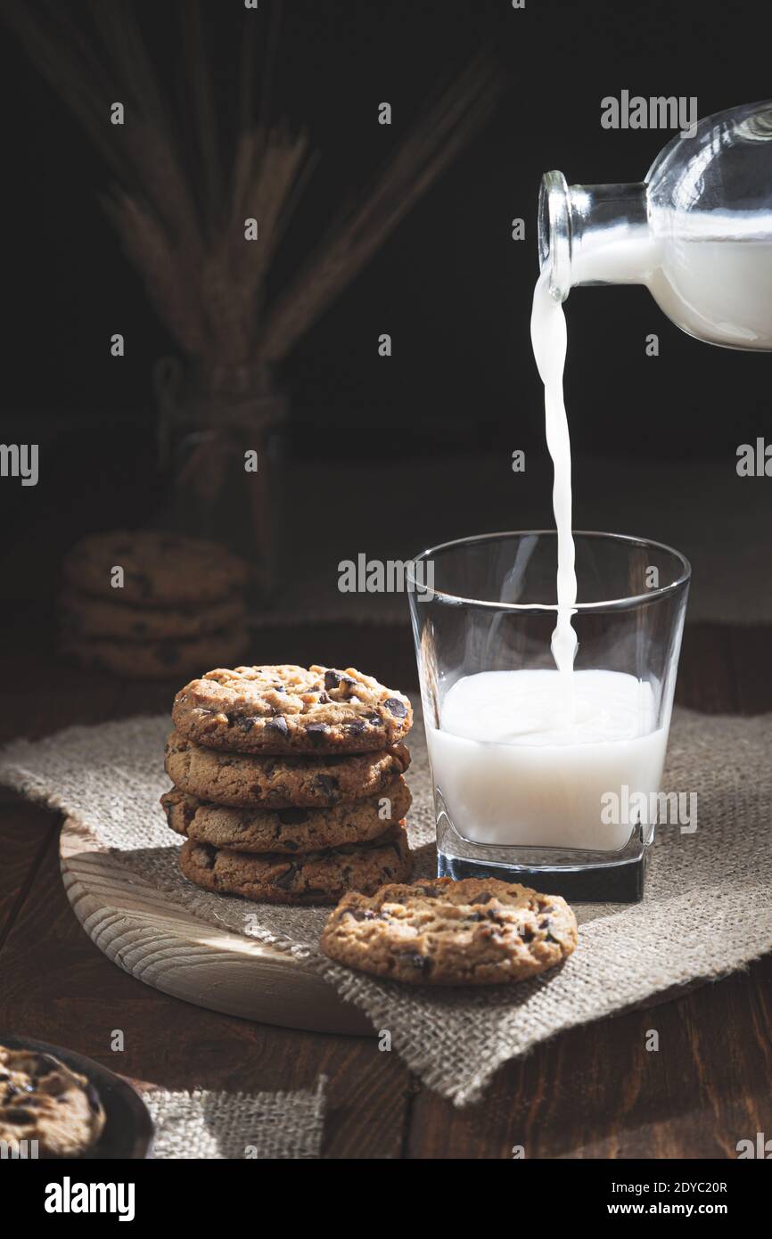 Chocolate Chip Cookies und Milchflasche verschüttet Milch in einem Glas auf einem Holzsockel, dunkler Hintergrund. Konzept für süße Speisen. Stockfoto