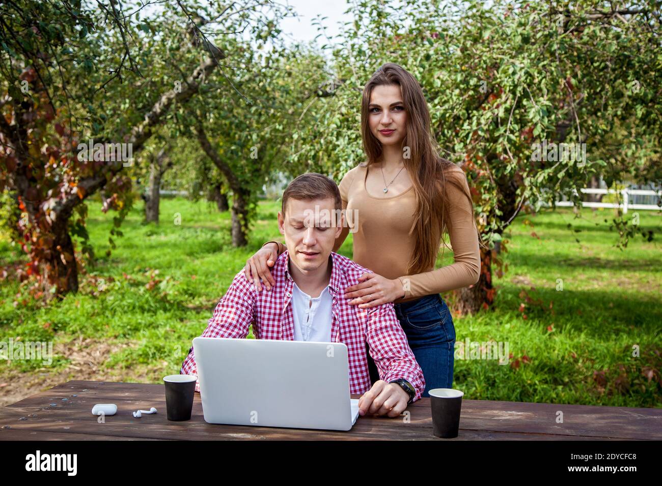 Lächelndes junges Paar verbringt Zeit zusammen im Park, studiert und arbeitet, während es mit einem Laptop an einem Holztisch sitzt. Kerl mit einem Mädchen mit Gadgets in Th Stockfoto