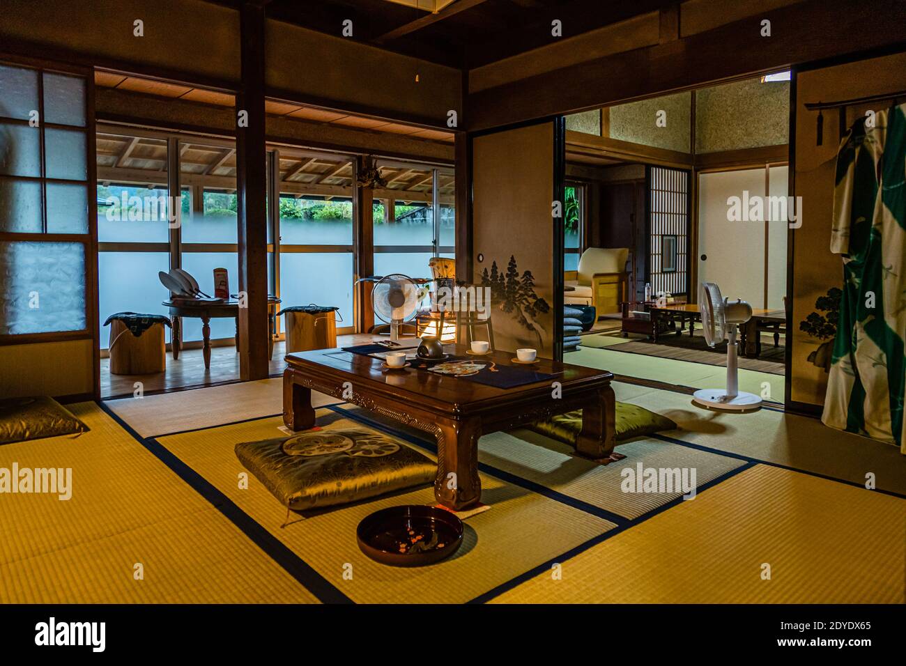 Ein gestreifter Yukata-Mantel hängt neben dem Eingang eines traditionellen Tatami-Zimmers mit Blick auf den Fluss in der Nähe von Kakegawa, Japan, zwei elektrische Ventilatoren kühlen den Sitzbereich am lackierten Tisch. Stockfoto