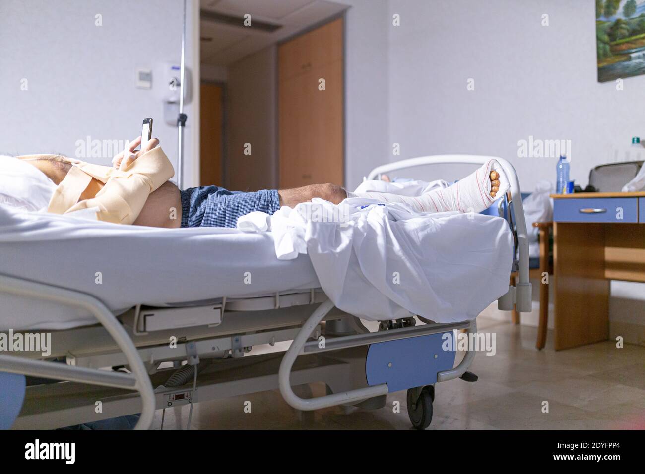 Patient liegt im Krankenhausbett mit gebrochenem Bein und schaut auf das Handy. Krankenhausaufenthalt und medizinisches Versorgungskonzept. Stockfoto