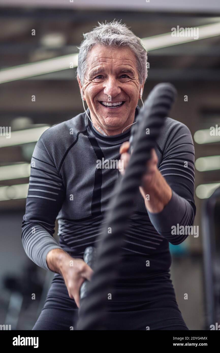 Der ältere Mann lacht beim Training mit Seilen im Fitnessstudio und trägt Kopfhörer. Stockfoto