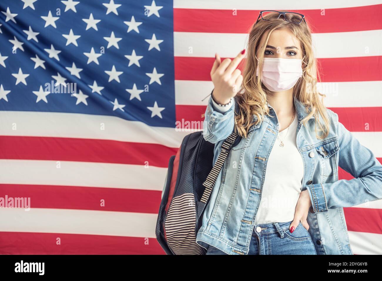 Junge blonde Studentin zeigt Mittelfinger-Geste zur Kamera, während sie vor der US-Flagge steht. Stockfoto